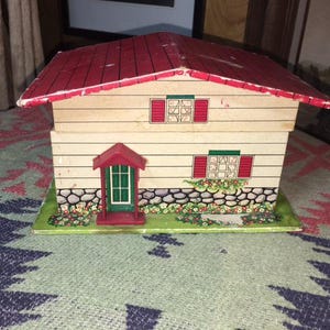 May include: A decorative wooden house with a red roof, beige walls, and green and red accents. The house features a small front porch, windows with shutters, and a stone and floral base. The house is a charming decorative item.