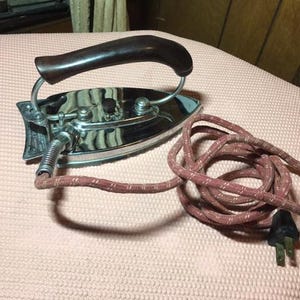 May include: Vintage chrome iron with a dark brown wooden handle. The iron has a silver body and a coiled cord with a pink and white pattern. The electrical plug is black. A classic appliance for ironing clothes.