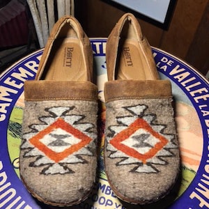 May include: A pair of brown suede slip-on shoes with a geometric pattern in orange, gray, and black on the front. The shoes have a felt-like texture and a brown leather trim. The brand name "Born" is visible on the insole.