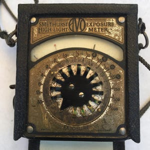 May include: Vintage black and gold Smethurst AVO Exposure Meter. The meter has a circular dial with numbers and a needle, and the text "SMETHURST HIGH-LIGHT" and "EXPOSURE METER" are visible. The meter is encased in a black leather case with a strap.