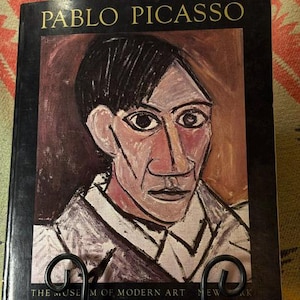May include: A hardcover book titled PABLO PICASSO THE MUSEUM OF MODERN ART NEW YORK featuring a portrait painting of a person with dark hair, a white collared shirt, and a brown background on the front cover, displayed on a black metal stand.