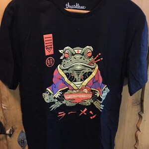 May include: Black t-shirt with a graphic of a frog wearing a headband and kimono, holding a bowl of ramen with chopsticks. The word "threadless" is printed on the collar. Japanese characters and the word "ramen" are printed on the shirt.