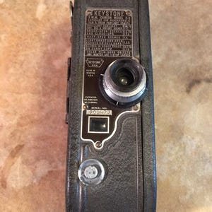 May include: A vintage Keystone M.M. camera, model X-8, in dark gray. The camera features a lens, dials, and a serial number plate. The text on the camera includes "KEYSTONE" and instructions for use. The camera is a classic piece of photography equipment.