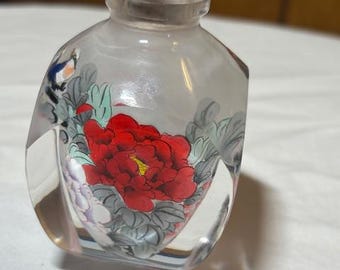 May include: A clear glass perfume bottle with a red-brown stopper. The bottle features a hand-painted design of a large red peony flower with green leaves and a small bird. The bottle has a unique, faceted shape.