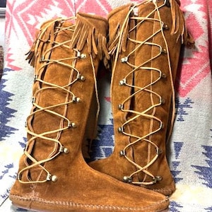 May include: A pair of knee-high brown suede boots featuring a lace-up front design with thin tan leather cords threaded through metal eyelets. The boots have a fringe detail along the top edge and a flat sole with visible stitching.