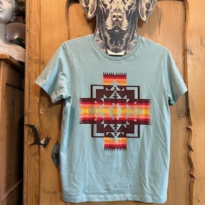 May include: A light blue t-shirt with a geometric design in shades of brown, orange, and yellow. The design is centered on the front of the shirt. The shirt is hanging on a wooden door.