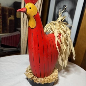 May include: A handcrafted wooden rooster figurine. The rooster is painted red with a yellow head and a red comb. It has a straw-like tail and a base with a straw-like trim. The figurine is a decorative item.