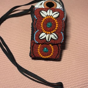May include: A small, rectangular beaded pouch with a black strap. The pouch is primarily red with intricate patterns of orange, white, and turquoise beads. The design includes floral and circular motifs, and the strap is a dark, woven material.