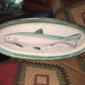 May include: An oval ceramic serving platter featuring a hand-painted fish illustration in shades of light blue, white, and grey. The platter has a wide, textured rim with a mottled green and brown finish, providing a rustic border to the central design.