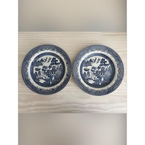 Puede incluir: Dos platos redondos de cerámica con un patrón de sauce en azul y blanco. El diseño representa un paisaje con pagodas, árboles y pájaros en vuelo, rodeado por un borde geométrico decorativo a lo largo del borde de cada plato.