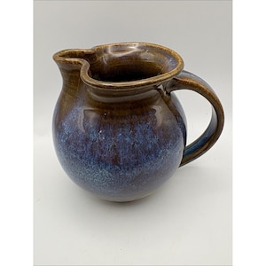 May include: A ceramic pitcher featuring a rounded body and a curved handle. The glaze transitions from a deep brown at the rim and handle to a mottled blue and purple finish across the lower section. It has a distinct spout with a wide opening.