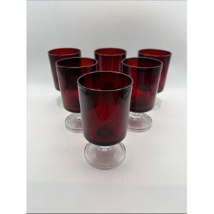 May include: A set of five matching drinking glasses featuring deep red bowls and clear glass stems and circular bases. The glasses are arranged in a triangular formation, showcasing a smooth, uniform design with a consistent color profile.