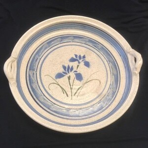 May include: A round, ceramic serving platter with a cream-colored base and blue accents. The platter features a hand-painted design of blue irises and green stems. Two handles are on opposite sides. The rim has a blue and white striped pattern.