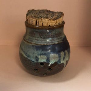 Puede incluir: Un tarro de ajo de cerámica hecho a mano con tapa de corcho. El tarro está esmaltado en tonos de azul, marrón y verde. Tiene varios agujeros pequeños cerca de la base. La tapa de corcho tiene una textura natural y rugosa.