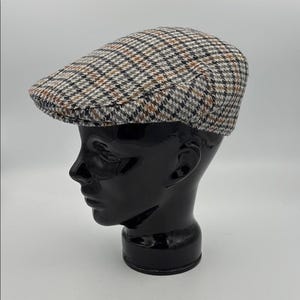 May include: A classic flat cap with a brown, black, and white plaid pattern. The cap is displayed on a black mannequin head. The cap has a rounded crown and a short, stiff brim. This style of hat is often associated with traditional or vintage fashion.
