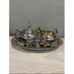 May include: A silver-plated tea service set consisting of a lidded sugar bowl and an open creamer, both featuring ornate handles and feet, arranged on a matching oval serving tray. The interior of the creamer has a gold-toned finish.