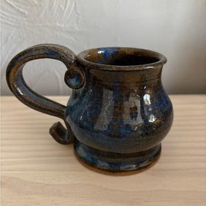 May include: A ceramic mug with a rounded body and a wide, curved handle featuring a small circular accent. The mug has a glossy glaze with blended tones of deep blue, dark brown, and hints of black, creating a mottled, variegated appearance.