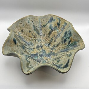 May include: A ceramic bowl with a wavy, scalloped rim and a speckled, mottled glaze in shades of beige, tan, deep blue, and forest green. The interior surface features a textured, organic pattern with blended colors and dark, irregular spots.