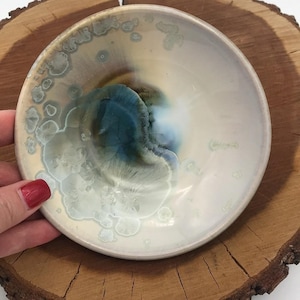May include: A small, round ceramic bowl with a unique glaze. The bowl features a creamy white base with blue and green crystalline patterns. The interior has a dark blue and brown swirl. The bowl is displayed on a wooden surface.