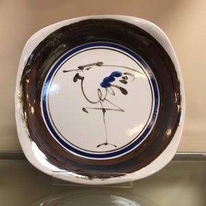 Pode incluir: Um prato de cerâmica quadrado com um centro branco com um desenho de pássaro azul e preto. A borda tem uma faixa marrom texturizada e uma faixa azul. A borda externa é branca.