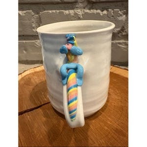 Könnte beinhalten: Ein weißer Keramikbecher mit einer texturierten, unebenen Oberfläche. Der Henkel ist als stilisierte Figur mit rosa Gesicht und blauen Gliedmaßen gestaltet, die an einem vertikalen Element mit diagonalen Streifen in Pastellgelb, Rosa, Blau und Grün sitzt.