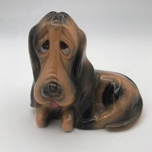 Puede incluir: Figura de cerámica de un perro Basset Hound en posición sentada. El perro tiene una expresión triste con ojos grandes y caídos y orejas largas. La figura es principalmente marrón y negra con un acabado brillante.