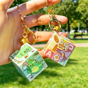 May include: Two keychains with gold-colored hardware and cube-shaped charms. One charm features a green dinosaur design with a rainbow, the other has colorful food and letter illustrations. The keychains are held up against a blurred green background.