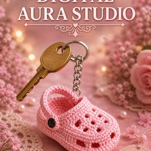May include: A pink crocheted baby shoe keychain with a gold key attached. The keychain is displayed on a pink surface with the text "DIGITAL AURA STUDIO" at the top. A tag reads "HANDMADE WITH LOVE".