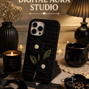 May include: A black phone case with a floral design. The case is made of a textured material and features a crocheted flower with white beaded petals and green leaves. The text "DIGITAL AURA STUDIO" is at the top.