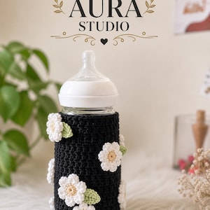 Könnte beinhalten: Ein Babyfläschchen mit weißem Sauger und einer schwarzen Häkelhülle. Die Hülle ist mit weißen und cremefarbenen Blumenapplikationen mit grünen Blättern verziert. Der Text "DIGITAL AURA STUDIO" steht oben.