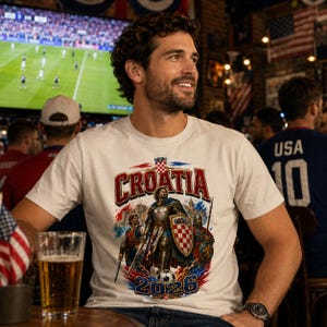 May include: White t-shirt with a Croatia soccer design. The graphic features the word "Croatia" in red and white, a knight, and the year "2026". The shirt is worn by a person in a bar setting, with a soccer game on a TV in the background.