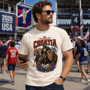 May include: Cream-colored t-shirt featuring the word "CROATIA" in red and white, with a graphic of a knight and soccer ball. The shirt also has the text "2026" and is set against a backdrop of red, white, and blue. The shirt is worn by a person.