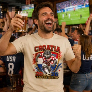 May include: A beige short-sleeve t-shirt featuring a graphic of a wolf wearing a red and white chequered football jersey. The design includes the text CROATIA, 2026, and SOCCER NATION, with a football and a shield emblem in red, white, and blue.