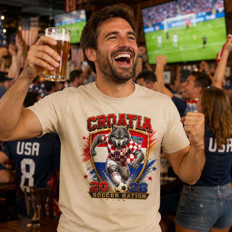 May include: A beige short-sleeve t-shirt featuring a graphic of a wolf wearing a red and white chequered football jersey. The design includes the text CROATIA, 2026, and SOCCER NATION, with a football and a shield emblem in red, white, and blue.