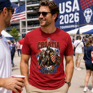 May include: A red t-shirt with a graphic featuring the word "Croatia" in gold lettering, a knight in armor, and the year "2026". The shirt is being worn by a person in sunglasses, with a crowd and an American flag in the background.