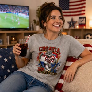 May include: A heather grey t-shirt featuring a graphic of a wolf wearing a red and white checkered football kit, kicking a football. The design includes the text Croatia, 2026, and Soccer Nation, set against a shield and star background.