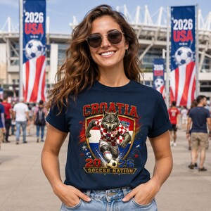 May include: A navy blue t-shirt featuring a graphic of a wolf wearing a red and white checkered football kit, running with a football. The text reads CROATIA 2026 SOCCER NATION, set against a shield background with red, white, and blue accents.