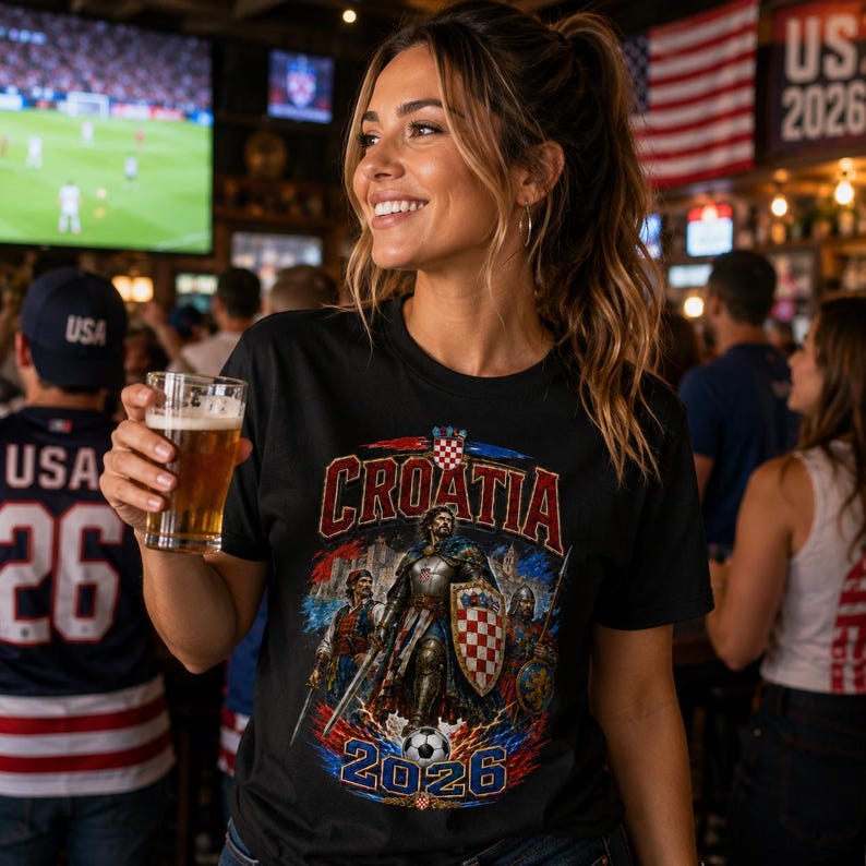 May include: Black t-shirt with a Croatia design featuring a knight, shield, and soccer ball. The shirt has the word "Croatia" in red and the year "2026" in blue. The background includes a bar setting with flags and people.