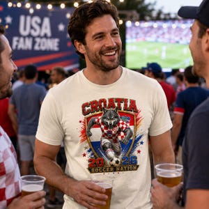 May include: A cream-coloured short-sleeve t-shirt featuring a graphic of a wolf wearing a red and white chequered soccer jersey. The design includes the text CROATIA, 2026, and SOCCER NATION, set against a shield background with red and blue accents.