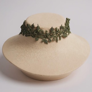 Peut inclure: Un collier ras du cou en dentelle verte avec un motif de feuilles et de vigne. Le collier est présenté sur une tête de mannequin de couleur crème. Le choker est fait d'une matière délicate et tissée.