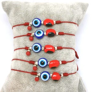 Op de afbeelding: Vijf rode draadarmbanden met blauwe evil eye-kralen en rode lieveheersbeestjeskralen. Elke armband heeft een kleine rode kraal die aan de knoop hangt. De armbanden worden gepresenteerd op een grijs kussen.