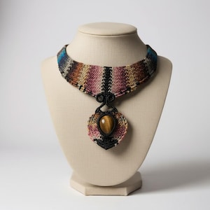 Puede incluir: Un collar artesanal con un collar ancho y tejido en tonos de rosa, naranja, marrón y azul. Un colgante negro y ornamentado sostiene una gran piedra ojo de tigre ovalada. El collar se exhibe en un busto de maniquí beige.