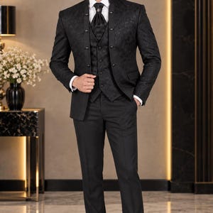 Könnte beinhalten: Ein schwarzer Dreiteiler bestehend aus einem Sakko mit Stehkragen und dezentem Ton-in-Ton-Blumenmuster, einer passenden Weste, einem weißen Hemd, einem schwarzen Plastron mit Ziernadel sowie einer schwarzen Anzughose und schwarzen Lederschuhen.