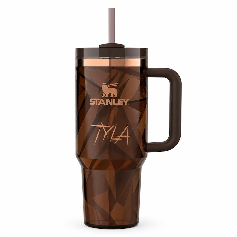 May include: A brown Stanley tumbler with a geometric pattern, handle, and straw. The tumbler features the Stanley logo and the name "Tyla" in a stylized font. The tumbler has a copper-colored band around the top.