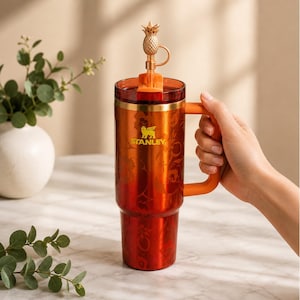 May include: A red and orange Stanley travel mug with a floral pattern and a pineapple-shaped stopper. The mug has an orange handle and a gold rim. The Stanley logo is visible on the front.