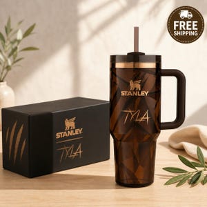 May include: A brown and gold Stanley tumbler with a handle and straw, next to a matching box. The tumbler has the Stanley logo and the name "TYLA" in gold. The box also features the Stanley logo and "TYLA".