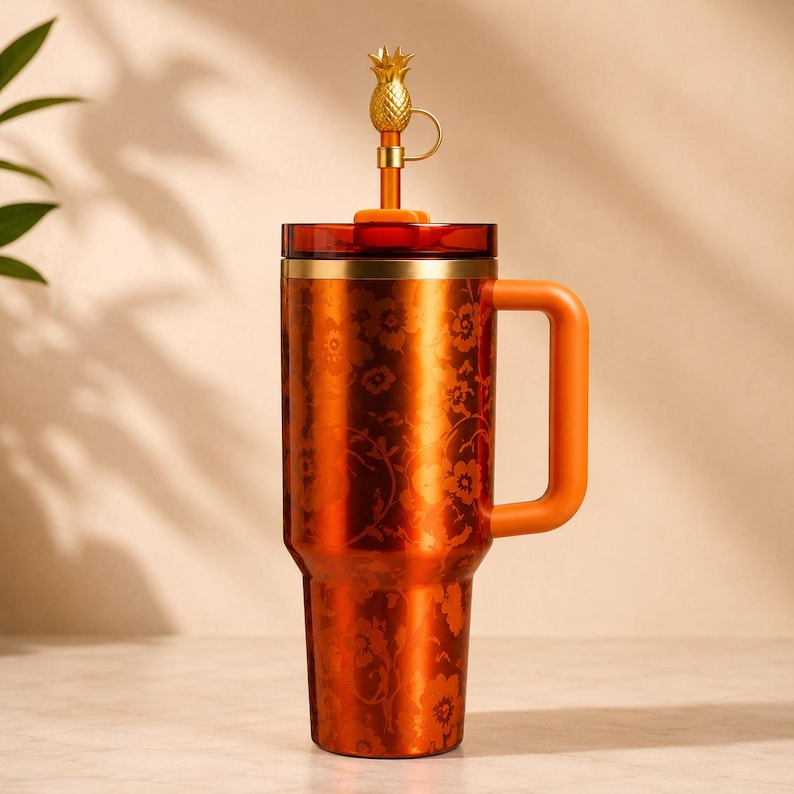 May include: An orange stainless steel travel mug with a floral pattern and a matching handle. The mug has a gold rim and a lid with a gold pineapple-shaped straw topper.