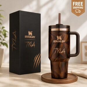 May include: A brown Stanley tumbler with a geometric pattern, a handle, and a straw, next to a black box with the Stanley logo and the word "TYLA". The tumbler sits on a wooden coaster.