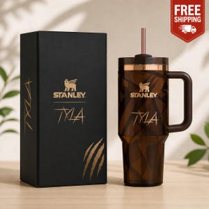May include: A brown Stanley tumbler with a handle and straw, next to a black box. The tumbler and box feature the Stanley logo and the word "TYLA" in gold. The tumbler has a gold band around the top.