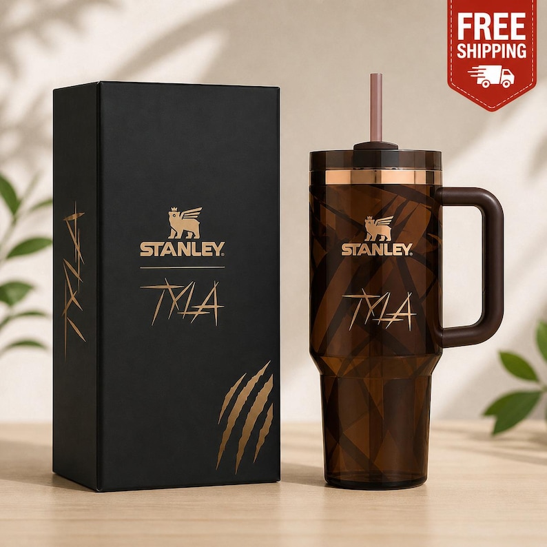 May include: A brown Stanley tumbler with a handle and straw, next to a black box. The tumbler and box feature the Stanley logo and the word "TYLA" in gold. The tumbler has a gold band around the top.